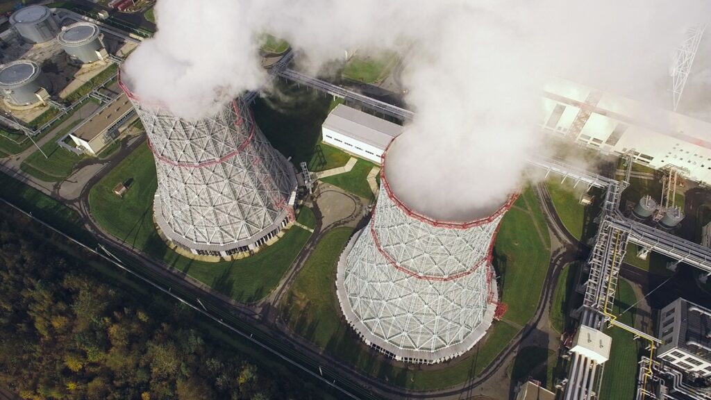 Aerial view of a nuclear power facility featuring two large cooling towers emitting steam, surrounded by industrial infrastructure, pipelines, and green landscaped grounds. The image represents the collaboration between Ontario and New York to expand nuclear energy development.