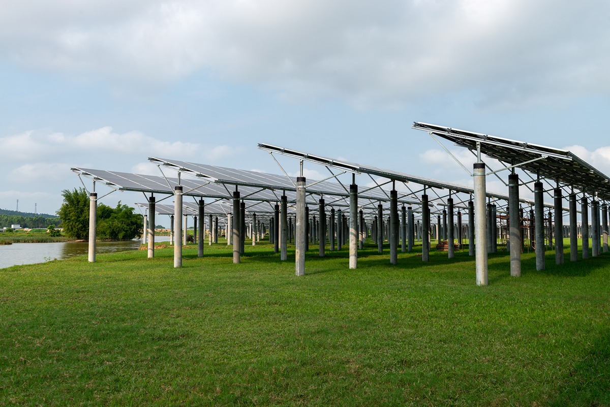 Rows of solar panel arrays mounted on elevated metal frames stretch across a grassy field near a waterway, part of a large-scale solar-storage project under development in California. The sky is partly cloudy, and the surrounding landscape includes trees and distant structures.