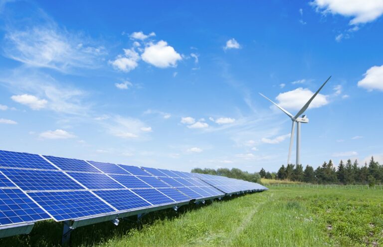 A solar farm with rows of blue solar panels in the foreground and a large white wind turbine in the background, set in a grassy field under a bright blue sky with scattered clouds.