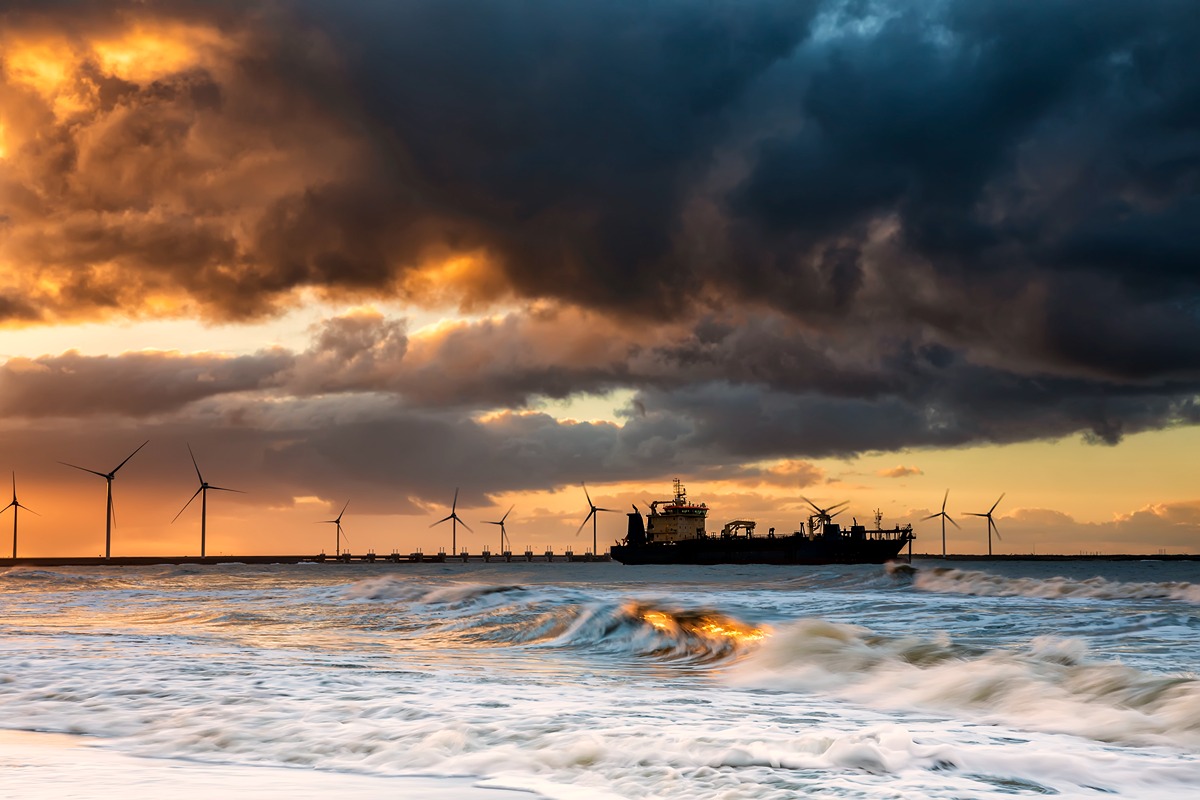 Dramatic coastal scene at sunset showing ocean waves, offshore wind turbines, and a large vessel silhouetted against a stormy sky. The image illustrates the potential of marine technology, like underwater cameras, to support the growth of America’s tidal energy industry.