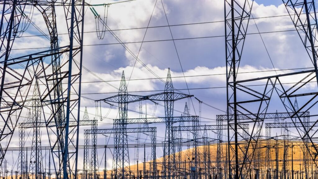 Dense network of electrical transmission towers and power lines set against a cloudy sky and rolling hills. The image emphasizes the scale and complexity of the U.S. power grid, relevant to discussions on rising energy demands from data centers.