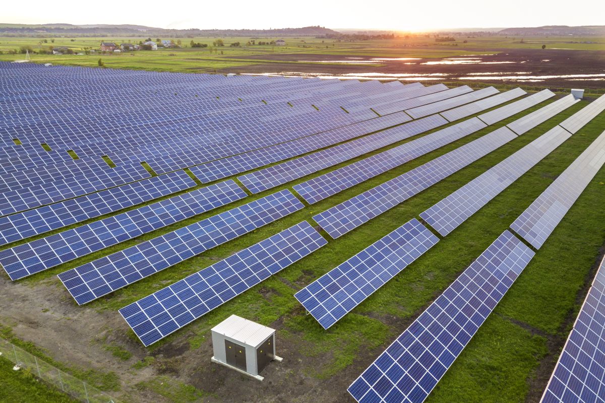 Aerial view of a large solar farm with long rows of blue solar panels stretching across green fields, with a small white utility structure in the foreground and the sun setting on the horizon.