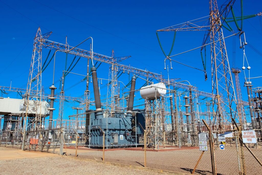 Electrical substation with large transformer units, metal lattice towers, and overhead power lines under a clear blue sky. Fenced utility infrastructure shows high voltage equipment and insulators used for power distribution.