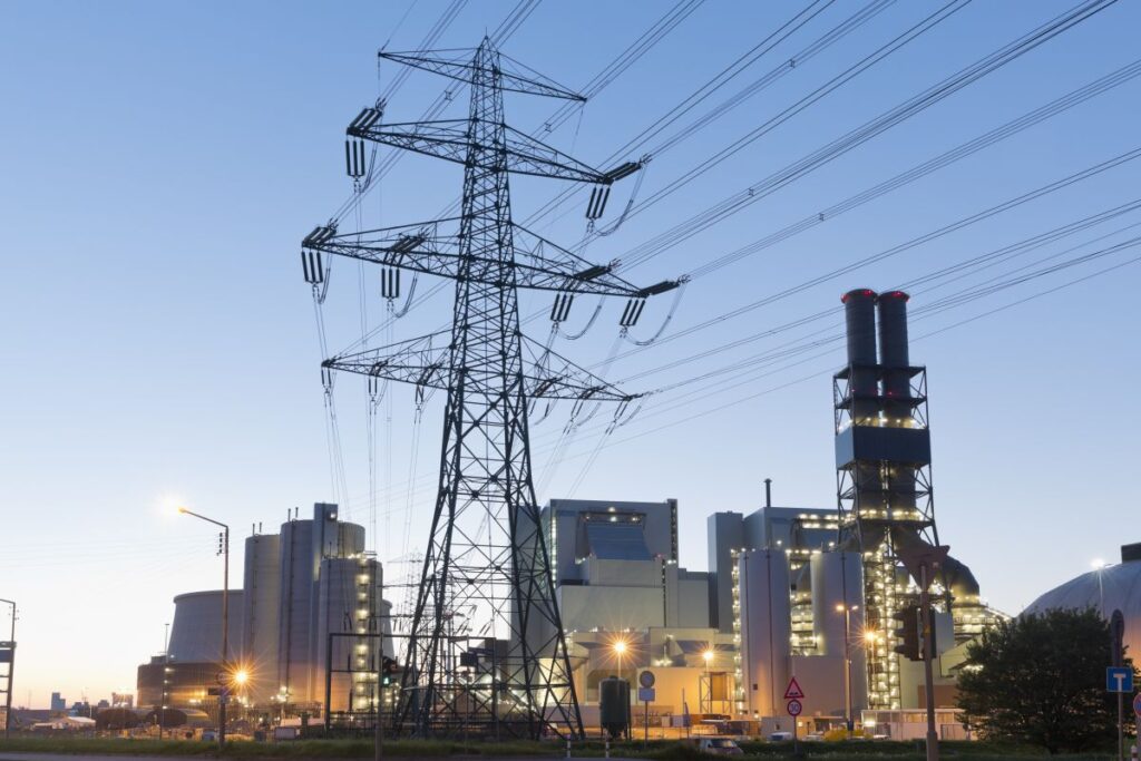 High voltage transmission tower and power lines stand in front of a large industrial power plant at dusk with lights glowing from the facility, illustrating large scale power generation and electricity distribution to the grid.