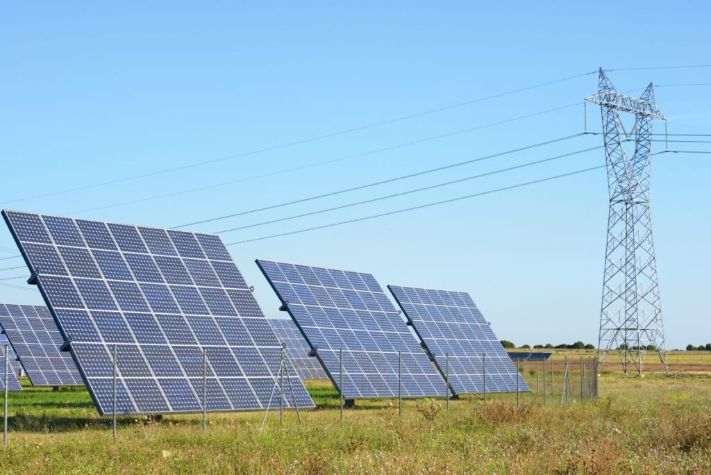 Solar panels in a grassy field beside a transmission tower and power lines showing renewable energy connected to the grid.