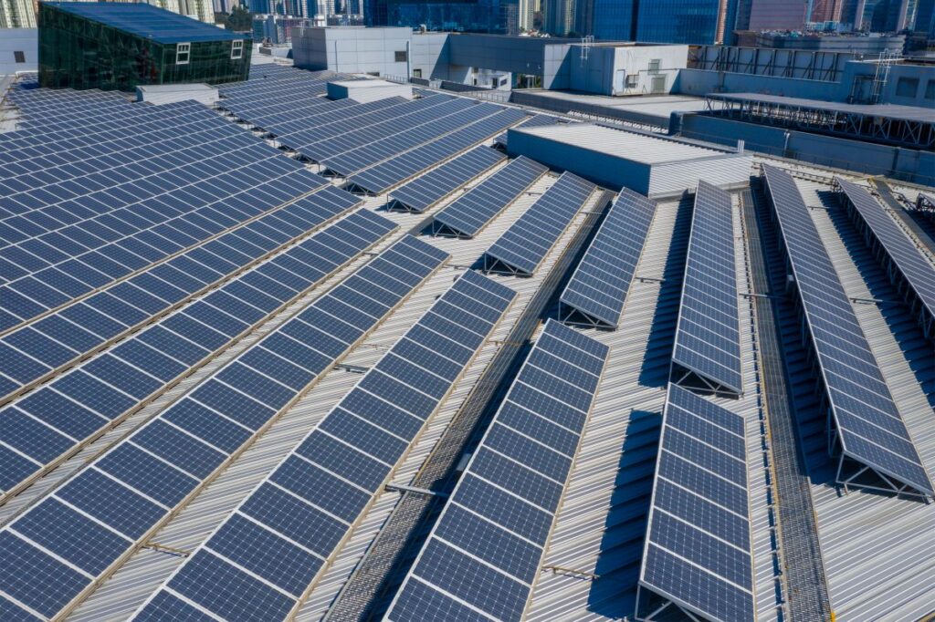 Aerial view of a large commercial rooftop covered in rows of blue solar panels installed on a metal building in a city. The panels are arranged in long angled lines across multiple roof sections, highlighting a large scale solar energy system in an urban environment.