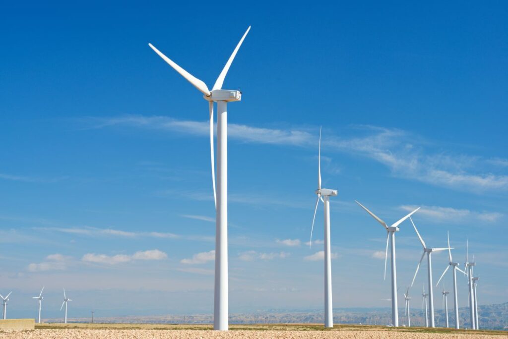 A row of tall white wind turbines stretches across a dry open field under a bright blue sky with scattered clouds, illustrating large scale wind energy production in a rural landscape.