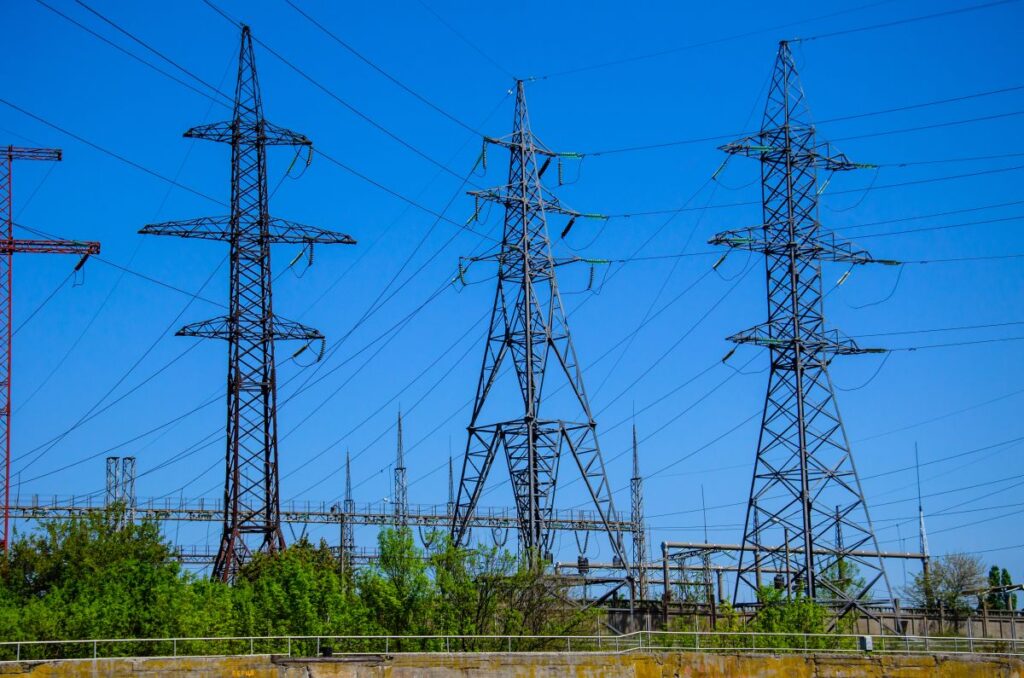 Tall metal electricity transmission towers connected by power lines stretch across a clear blue sky above a green field. The image highlights large scale energy infrastructure used to distribute electricity across long distances.