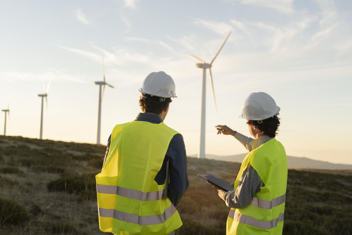 Two workers in safety gear review wind turbines at sunset, pointing toward a large-scale offshore wind project.