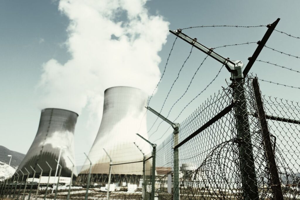 Cooling towers and industrial structures at a nuclear power plant rise behind a barbed wire fence under a cloudy sky, illustrating NRC issues 100th renewed license milestone for nuclear facility operations.