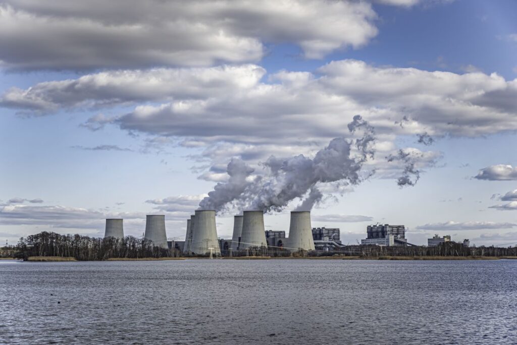 Wide view of a nuclear power plant with multiple cooling towers releasing white steam across a river under a cloudy sky, representing NRC issues 100th renewed license milestone for continued plant operation.