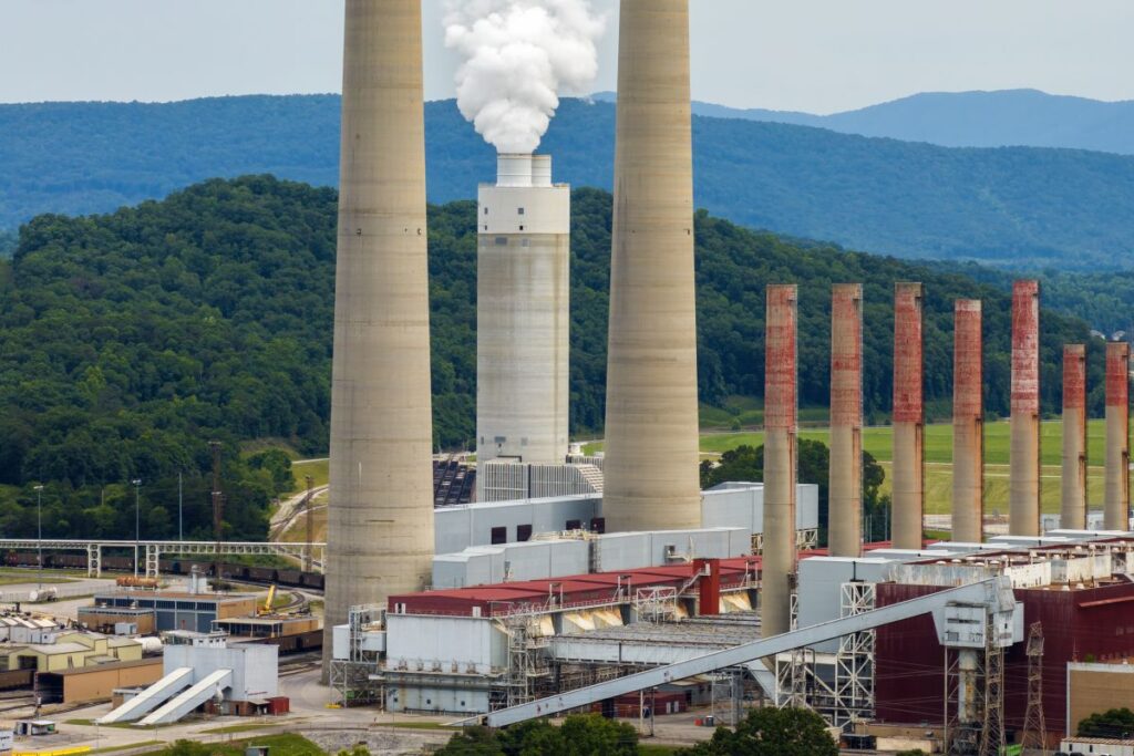 Coal-fired power plant producing electricity Fossil fuel usage for energy production A coal plant with two tall smokestacks and an industrial facility in front of forested hills. White steam rises from the central stack while older rust colored stacks stand nearby.