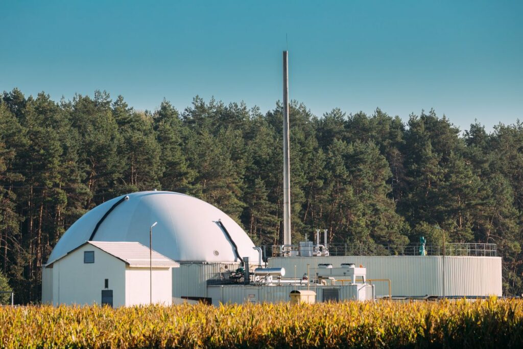 Industrial facility with a large white geodesic dome, tall chimney, and low white buildings near a cornfield.