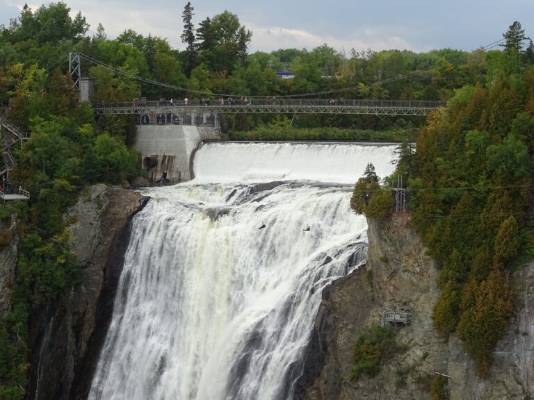 Bridge over a large waterfall with people walking along the railing, surrounded by forest.