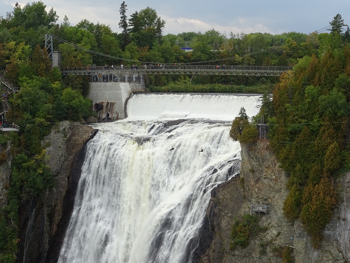 Bridge over a large waterfall with people walking along the railing, surrounded by forest.