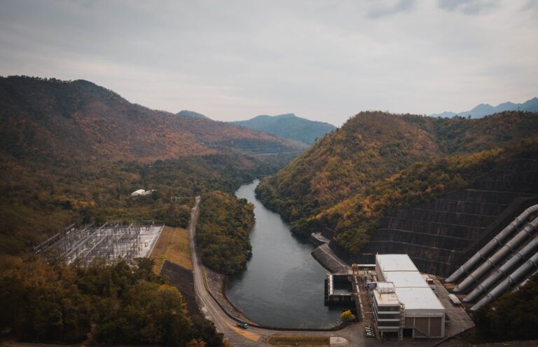 Di-Hydro Research Project Tackles Digitalization of Hydropower Plants Through Sensor Development-Certrec A winding river runs through a valley flanked by forested hills, with a dam and industrial buildings on the right and a power substation on the left.
