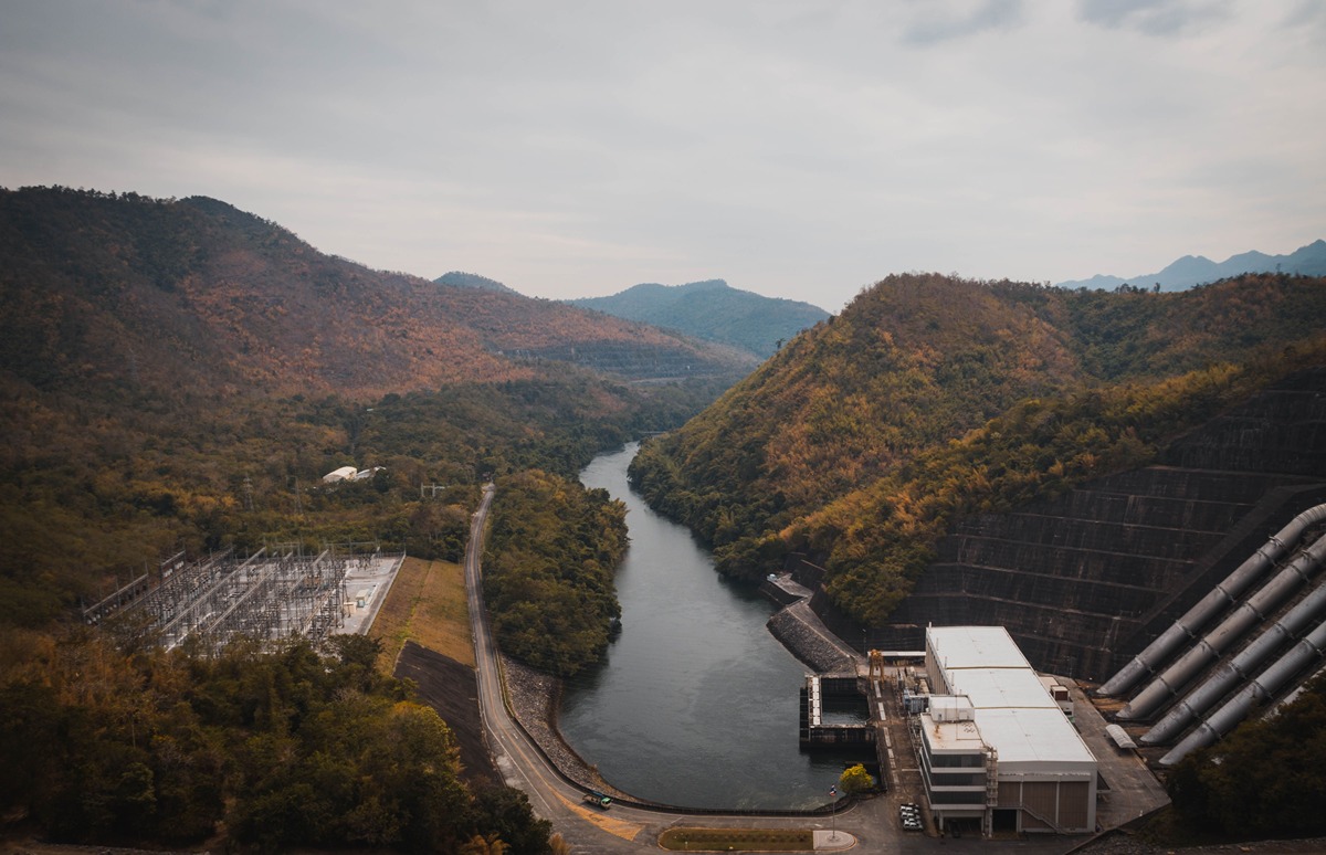 A winding river runs through a valley flanked by forested hills, with a dam and industrial buildings on the right and a power substation on the left.