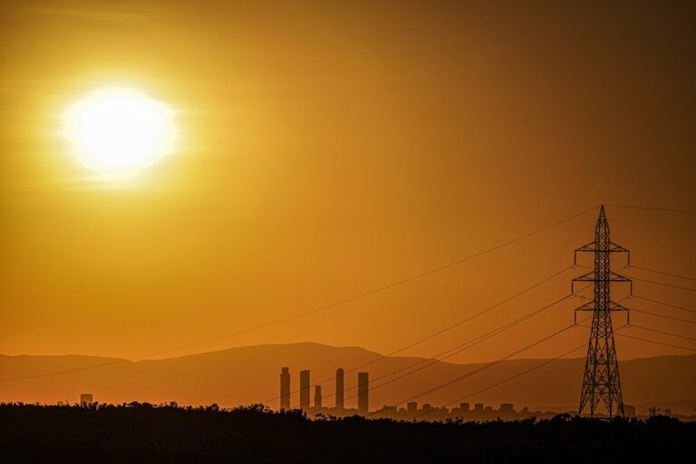 Energy Secretary Secures Grid Across 17 States amid Period of Hot Weather-Certrec Orange sunset over a distant industrial skyline with silhouetted power lines and a lattice transmission tower in the foreground.