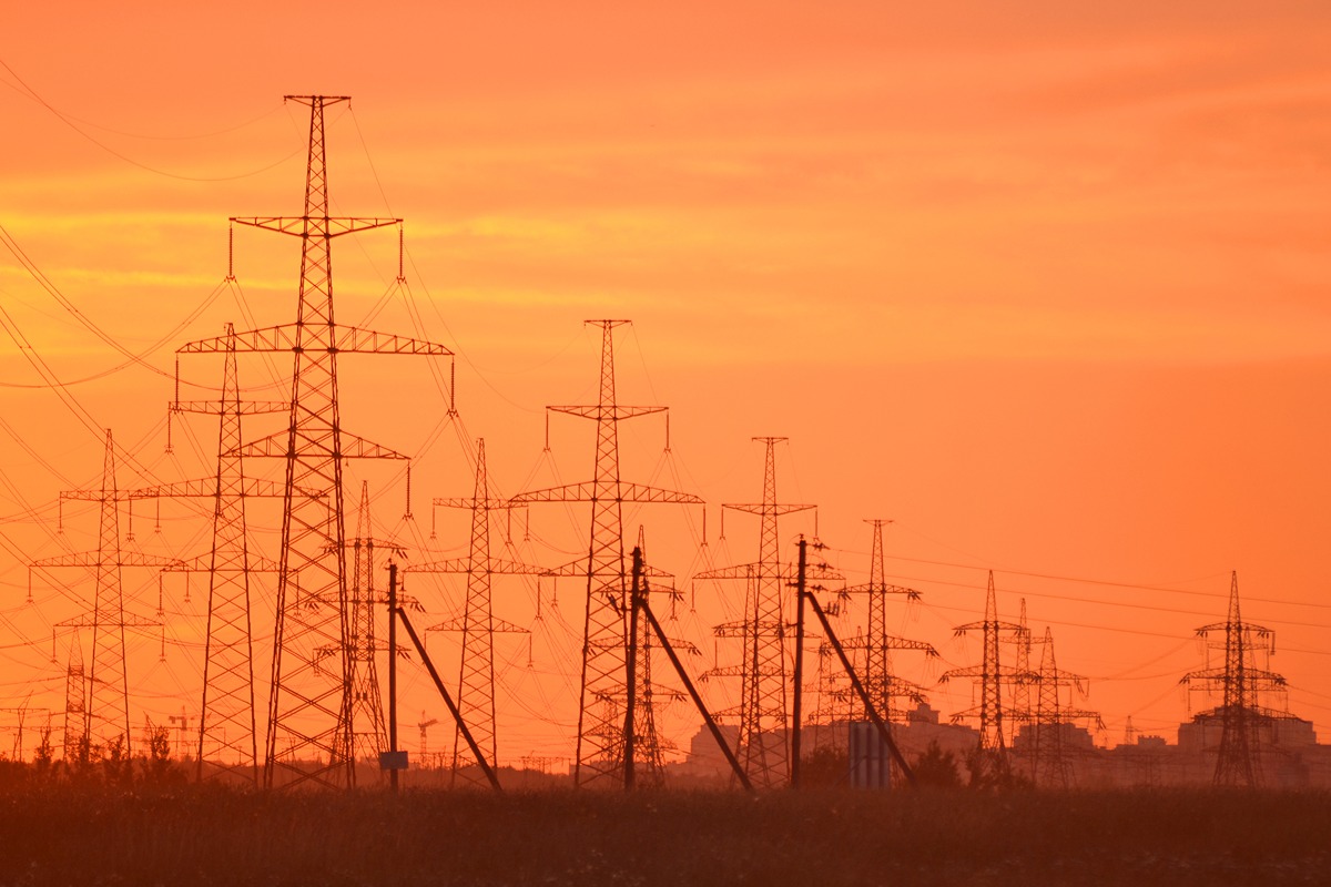 Silhouettes of tall transmission towers and power lines at sunset over a flat field.