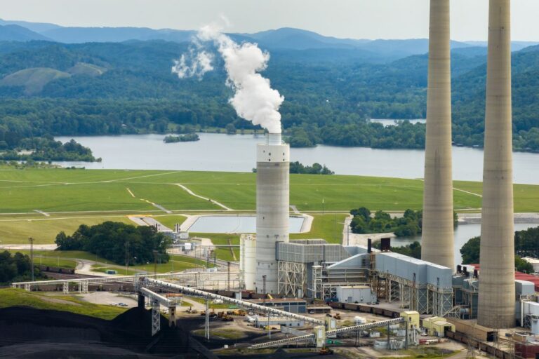 Industrial power plant with a tall smokestack emitting steam, adjacent cooling towers, and green hills in the background by a lake.