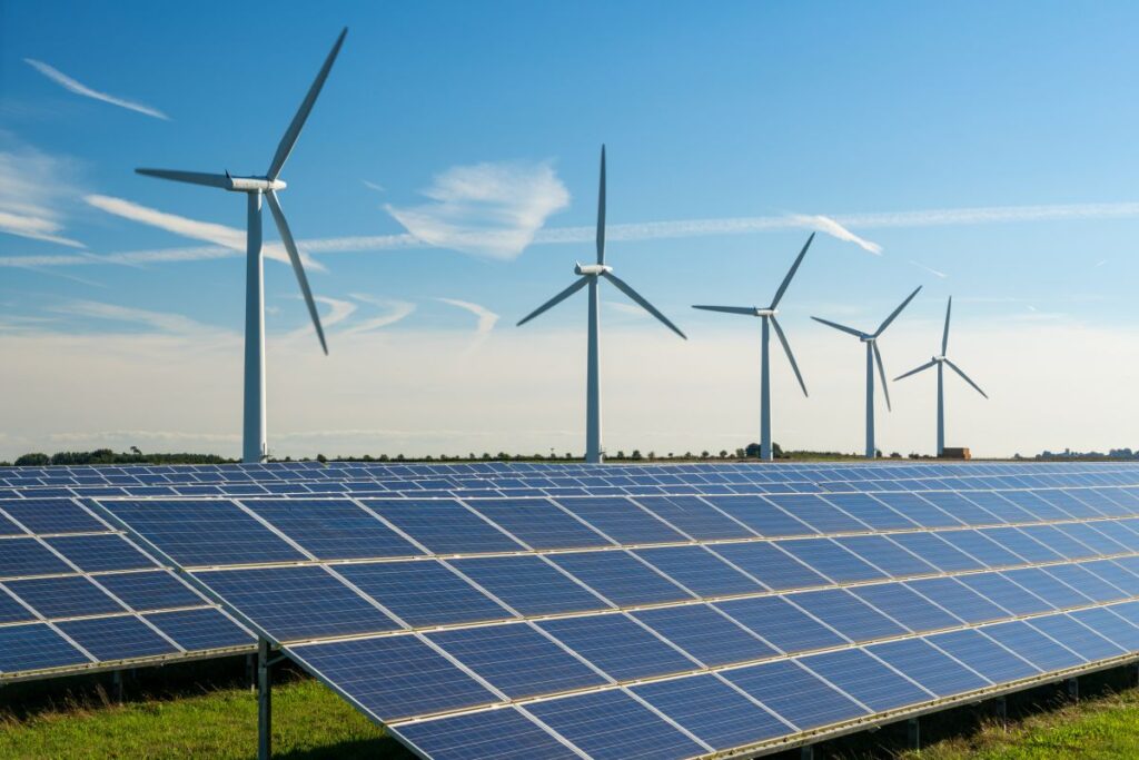 Solar panels in the foreground with wind turbines on a clear sunny day in the background, illustrating renewable energy.