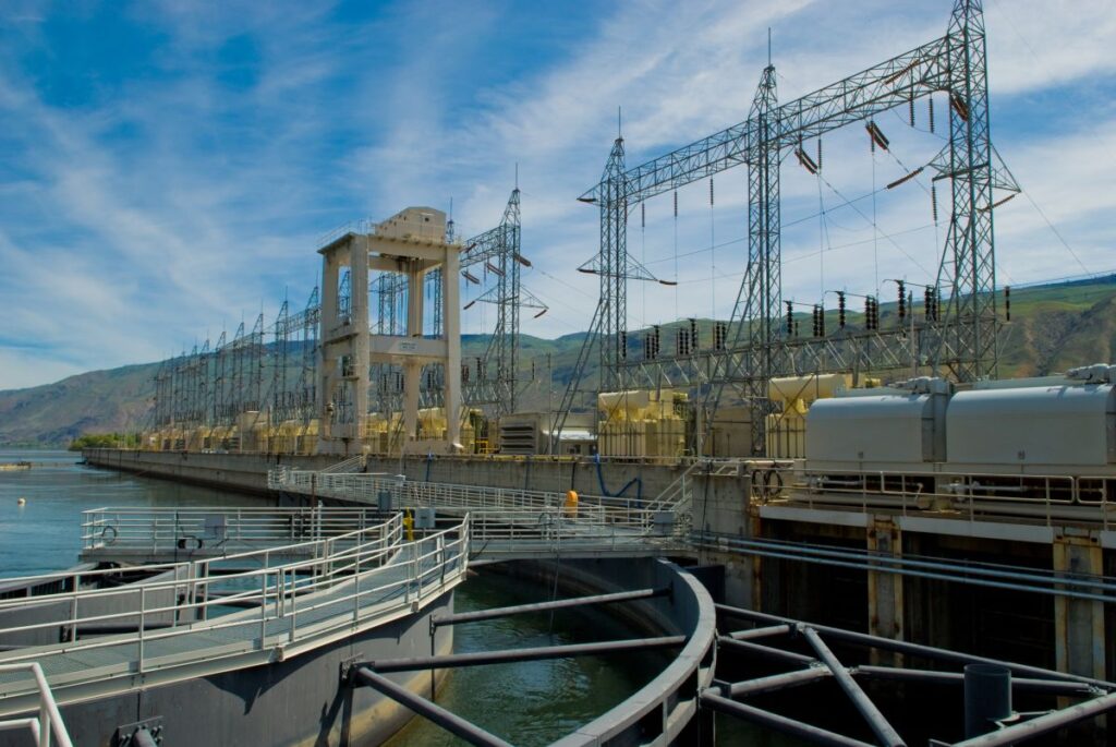 Hydroelectric dam facility with high-voltage towers and spillways along a waterway under a blue sky.