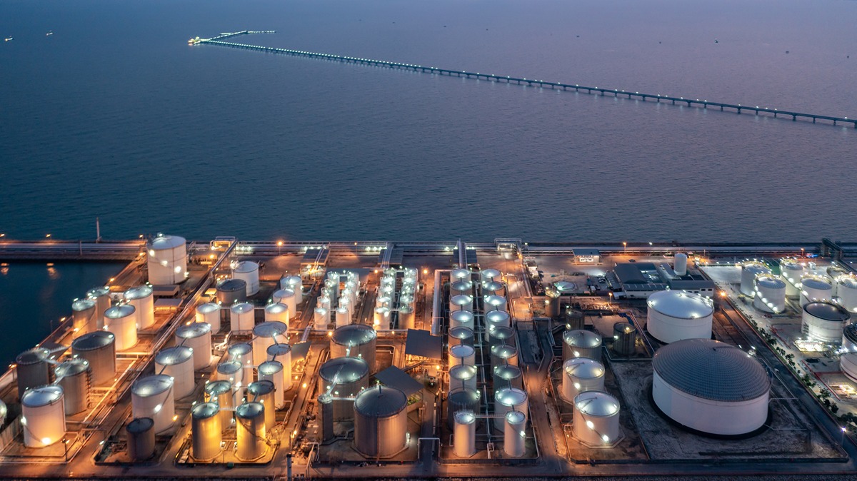 Aerial view of a coastal oil refinery with numerous white storage tanks and a long pier extending into the sea at dusk.