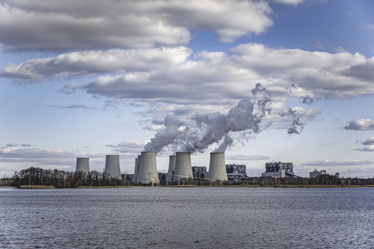 Industrial power plant along a river with several large cooling towers emitting steam under a cloudy sky.