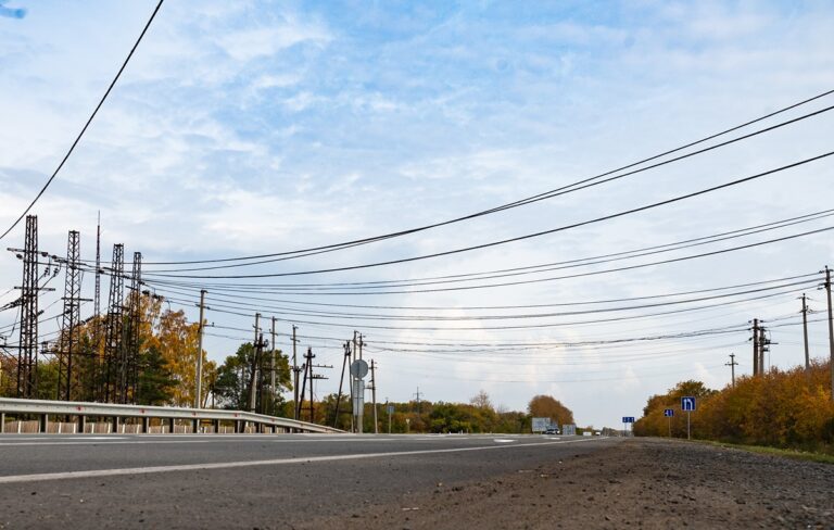 Long road lined with numerous overhead power lines and autumn trees under a blue sky, viewed from ground level.
