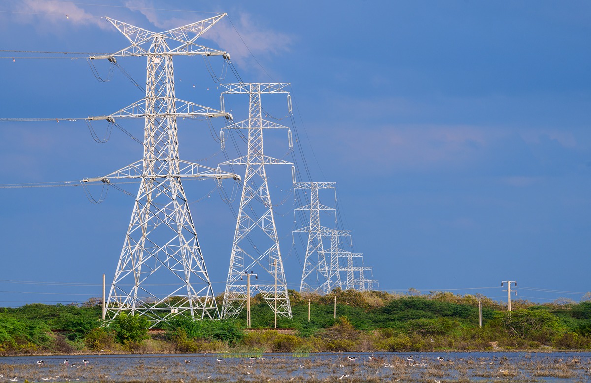 Row of tall metal electricity transmission towers with power lines stretching across a blue sky over a grassy landscape.
