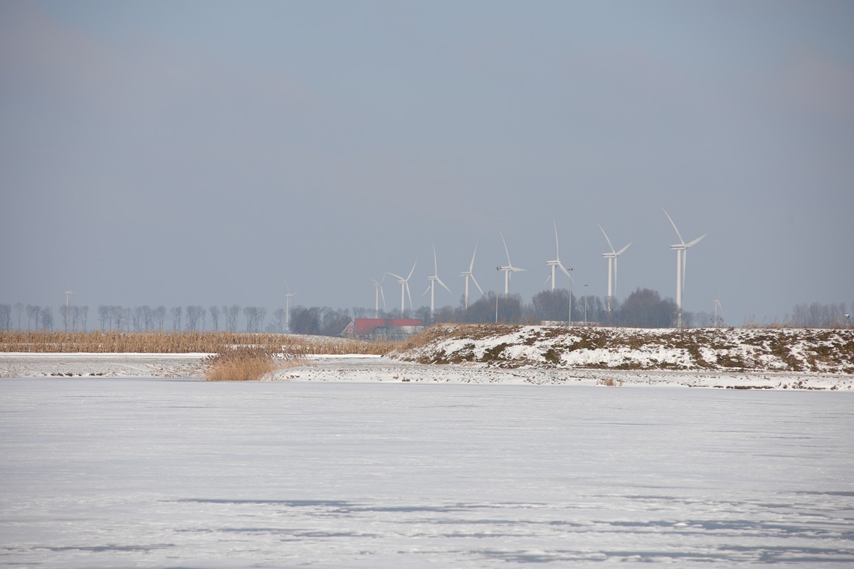 Snowy landscape with a row of wind turbines on the horizon and a red-roofed barn among trees in the distance.